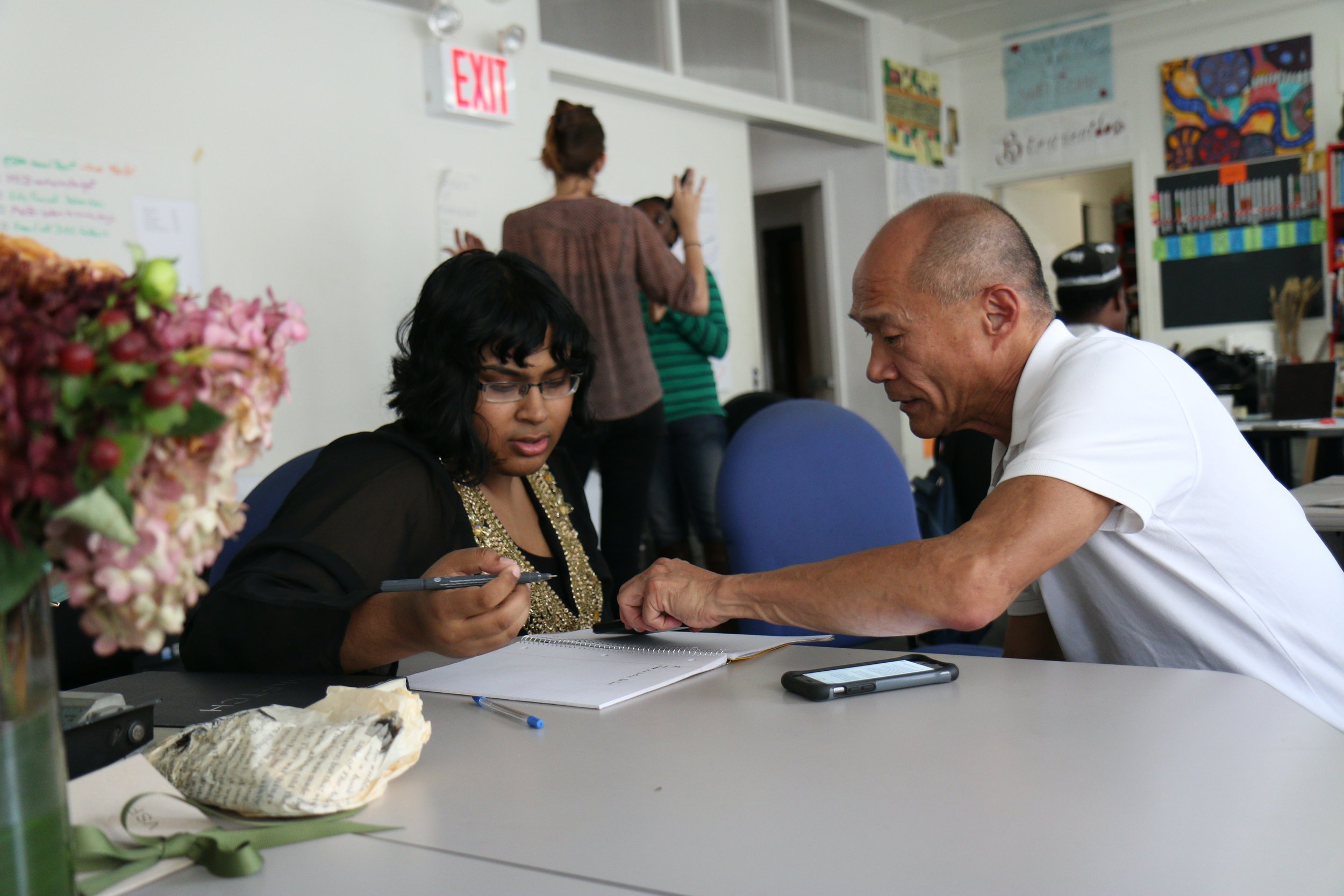 An older man and younger woman sit at a table both looking at a notebook. He is explaining something and pointing it out on the paper. They are both people of color.