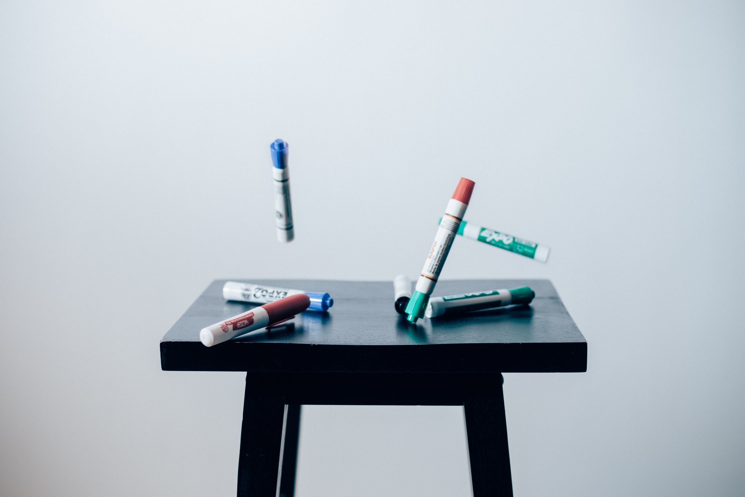 A photo of a small black stool with different colored dry erase markers falling around it