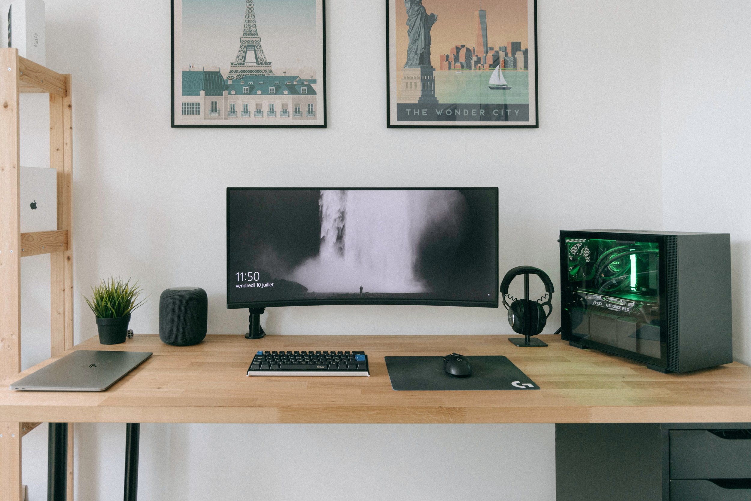Photo of a home office work space consisting of a computer, speaker, headset, keyboard, mouse, and posters of New York and Paris on the wall