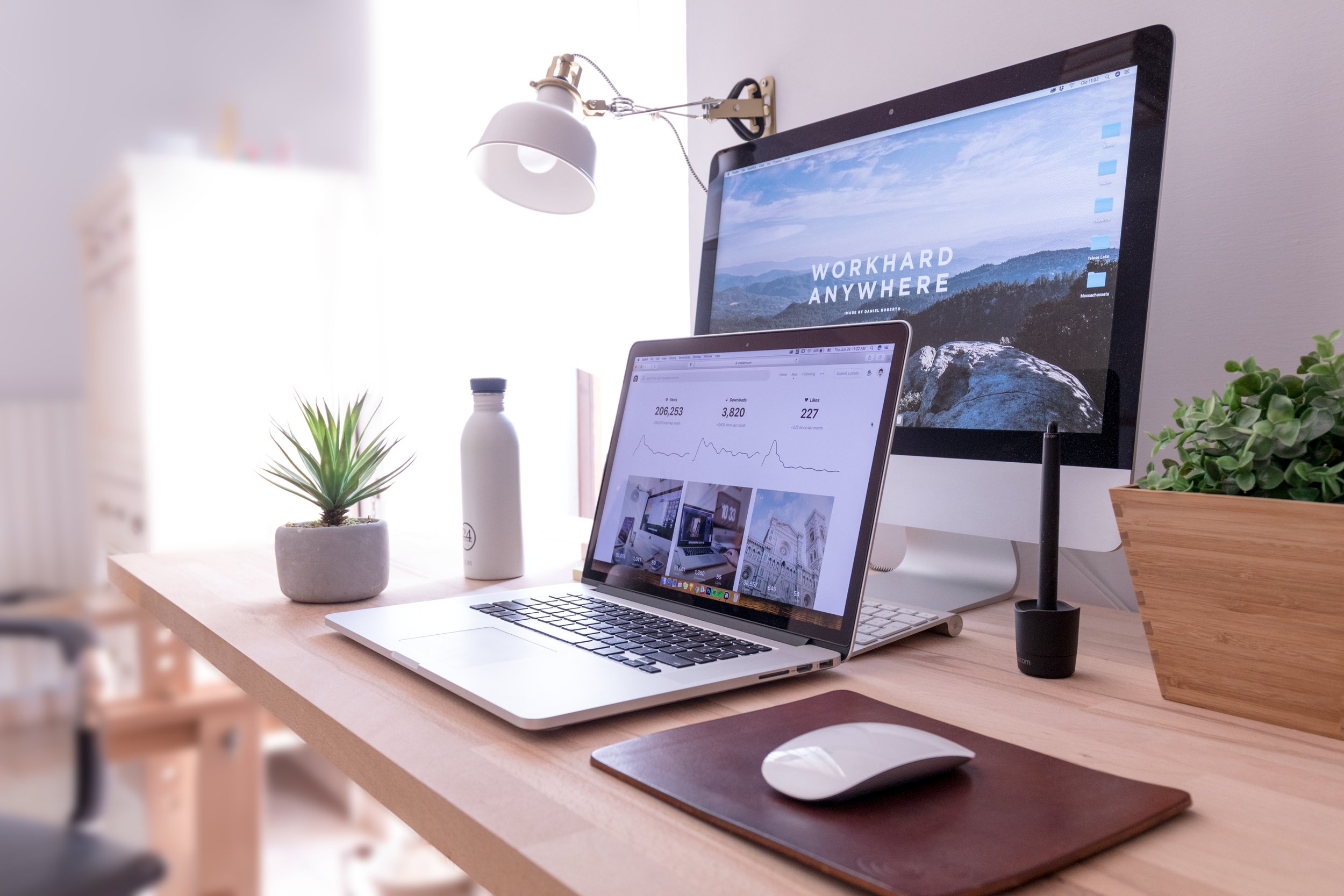 Photo of a desk in a home office with a desktop monitor and laptop, as well as reading lamp and house plant