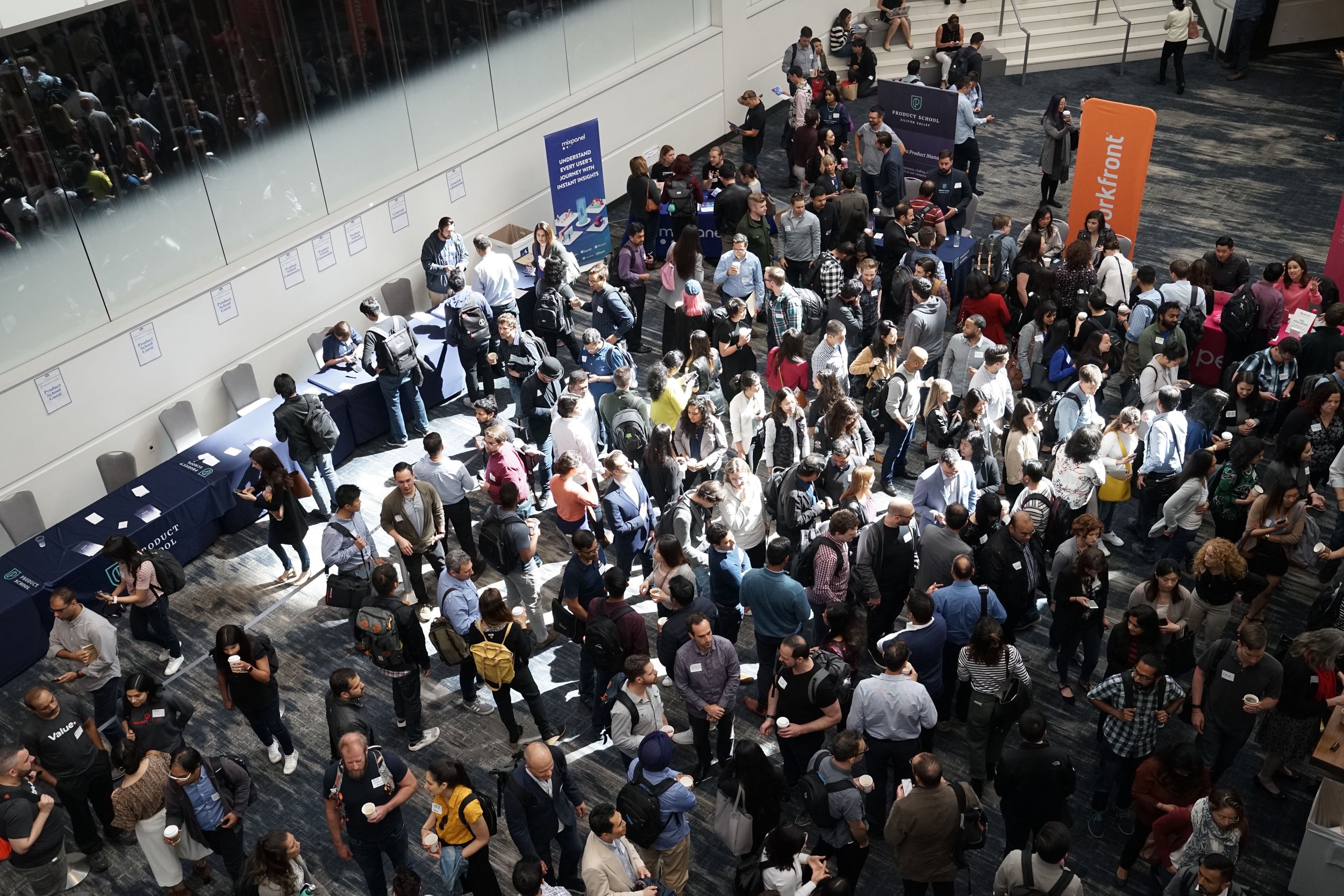 Overhead photo of a crowd at a jobs fair. A hundred or so diverse people in business outfits mingle in a conference hall.