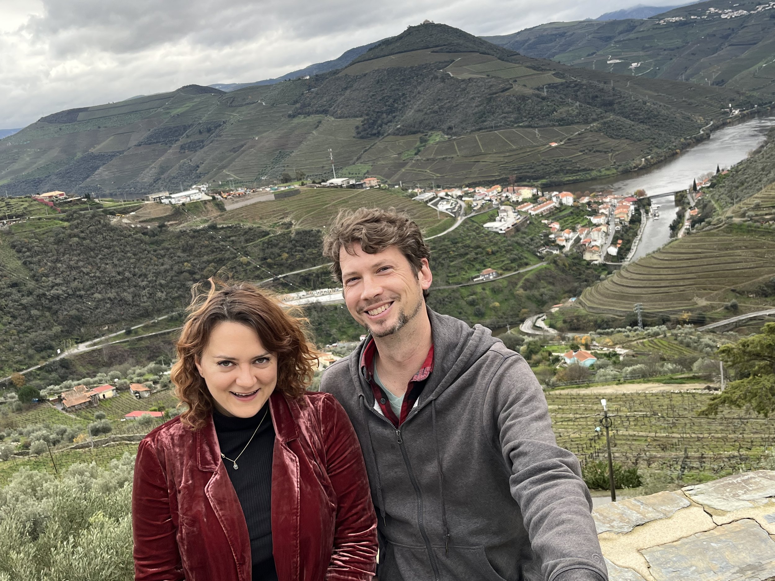 Photo of a smiling white man and woman with mountains, a river, and a village in Portugal in the background.