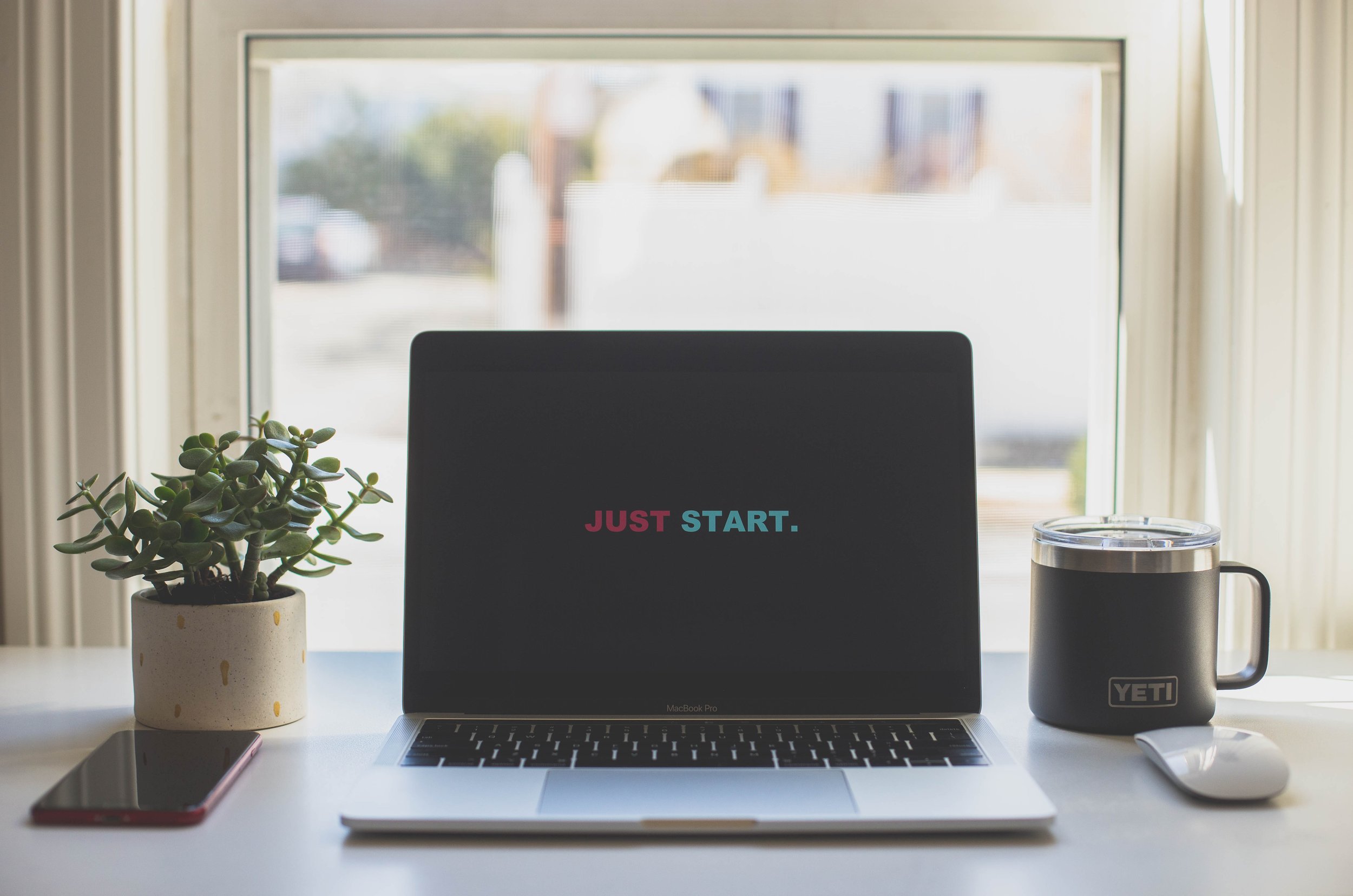 Photo of a desk in a home office with a house plant, YETI coffee tumbler, and open laptop that says "JUST START" on the screen