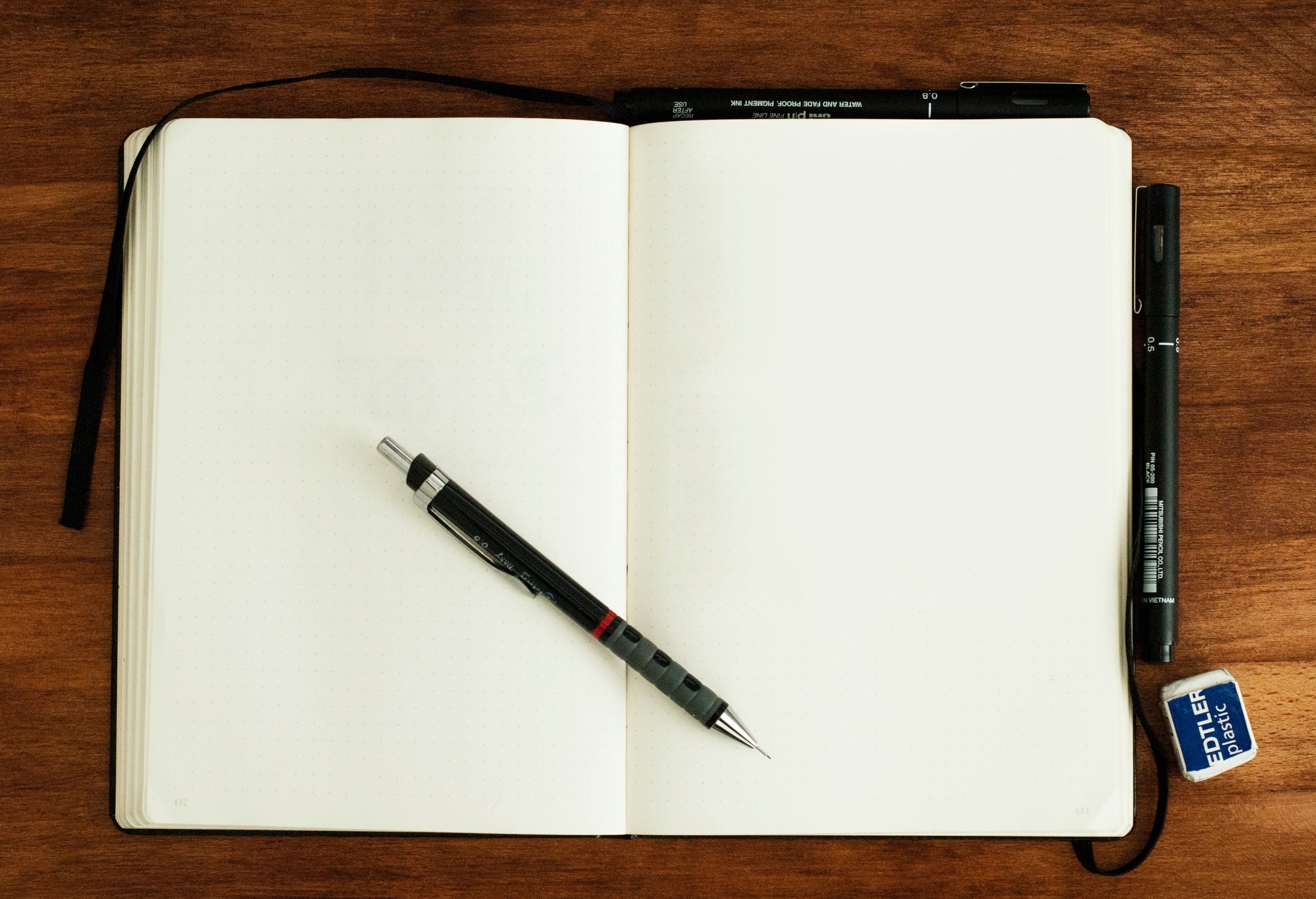Photo of a notebook open to a blank page laying on a wooden table with a pen on top.