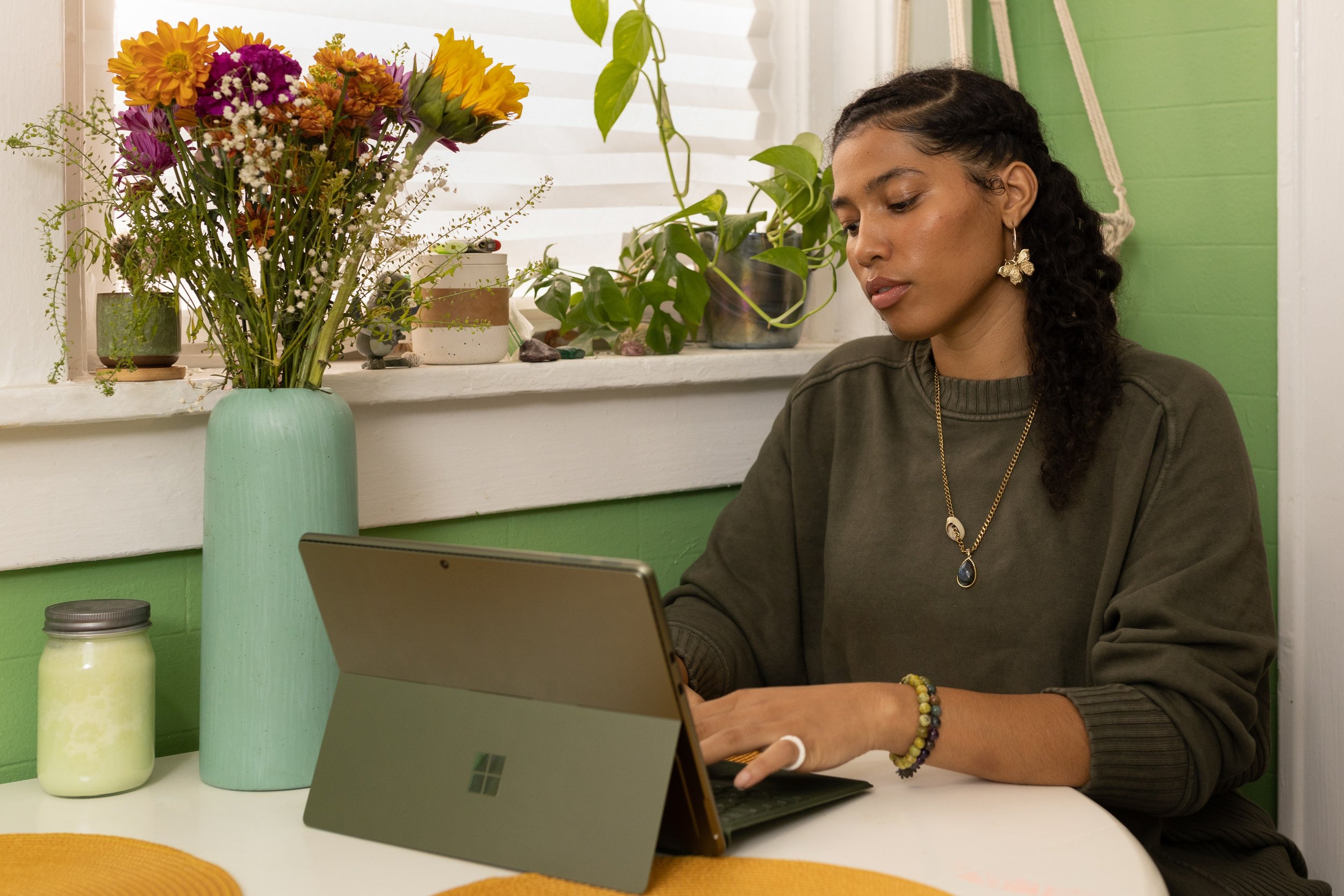 Photo of a young mixed race woman typing on her computer. She is sitting in a trendy space with green walls, macrame, and pots of plants.