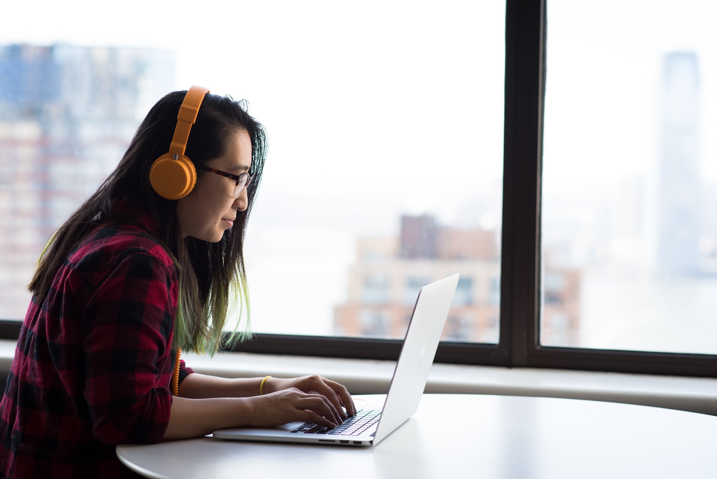 Photo of a young Asian woman with green tips in her hair and a bright orange headset working on her laptop in front of a large window with a city view