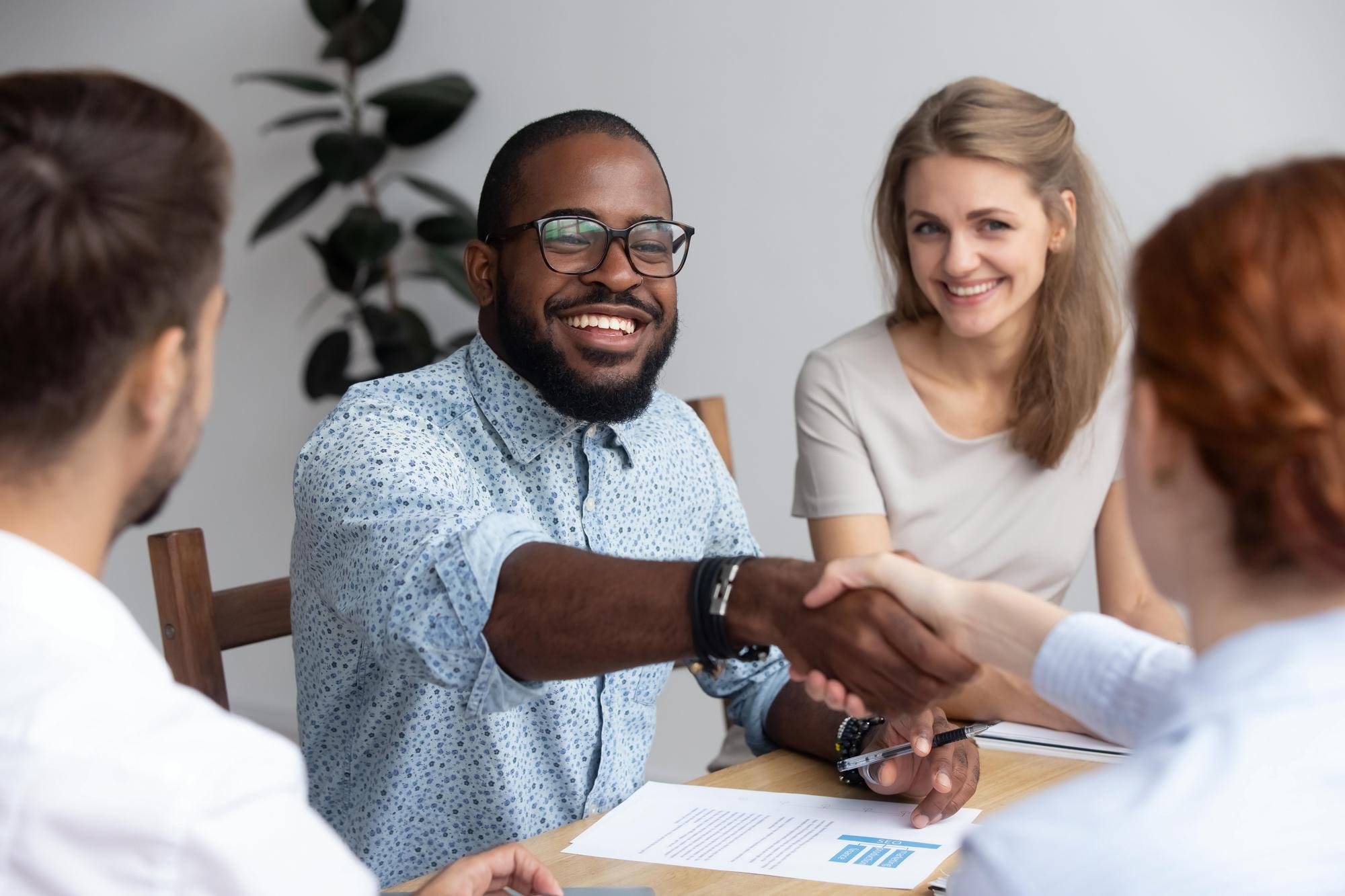 A young Black businessman shakes hand with someone over a stack of formal looking documents