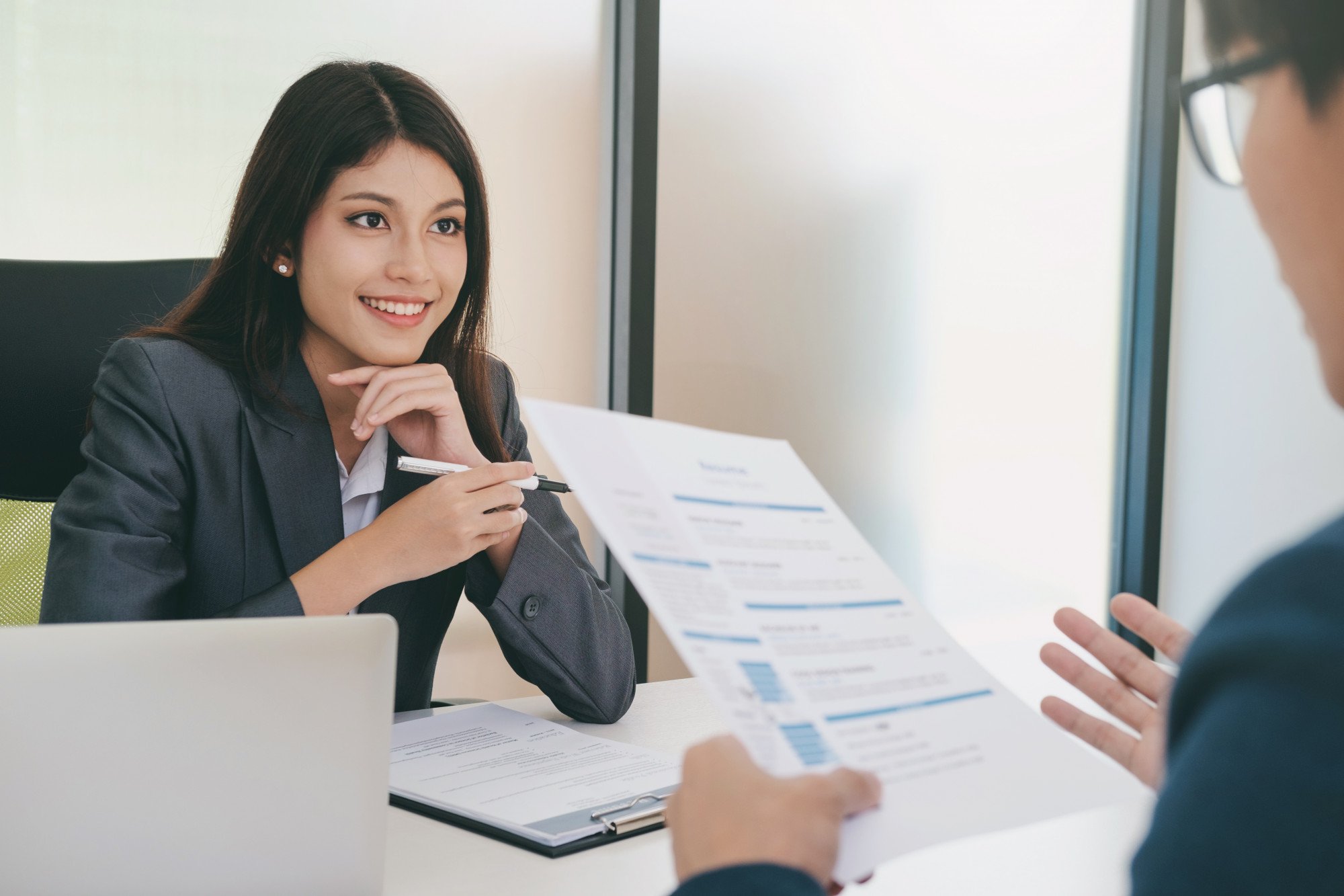 Two young Asian businesspeople participating in an interview as the man looks over a resume and the woman smiles while responding to a question