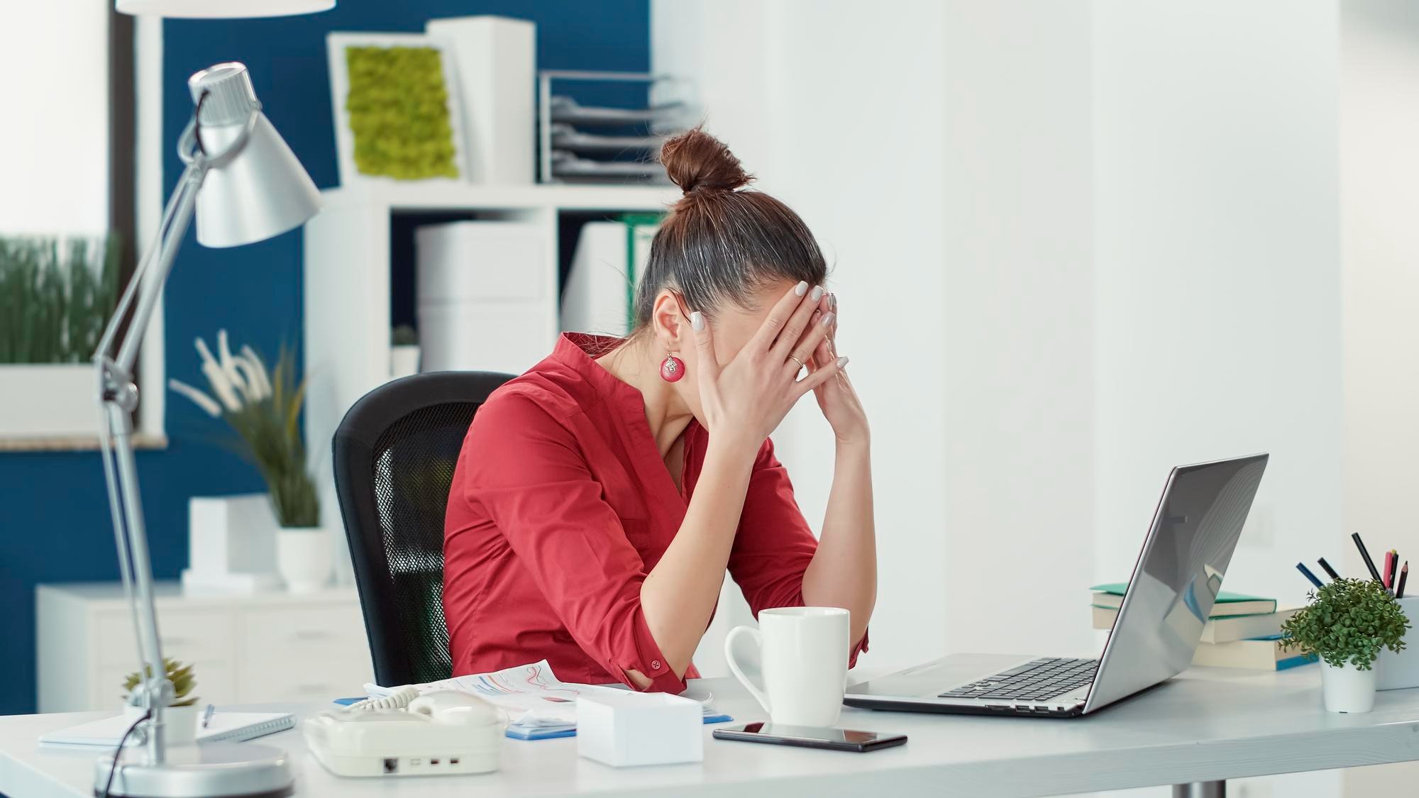 A female office worker sitting in front of her computer sits stressed with her hands on her face.