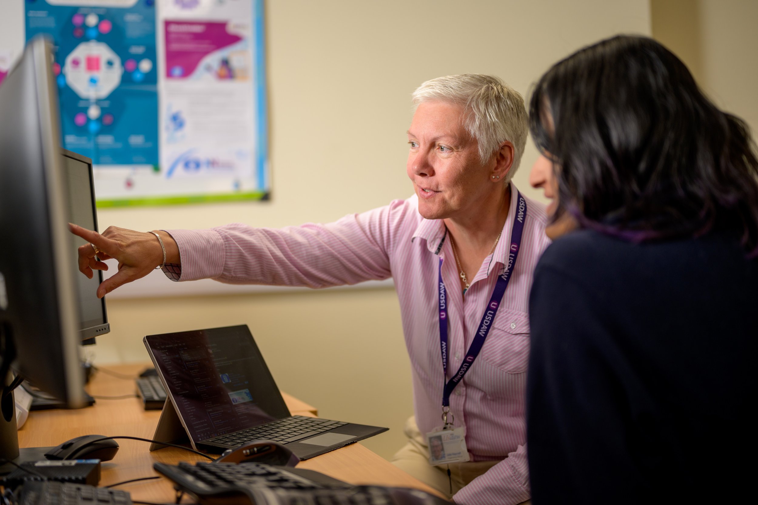 An older woman in an office points out something on a computer screen to a younger woman