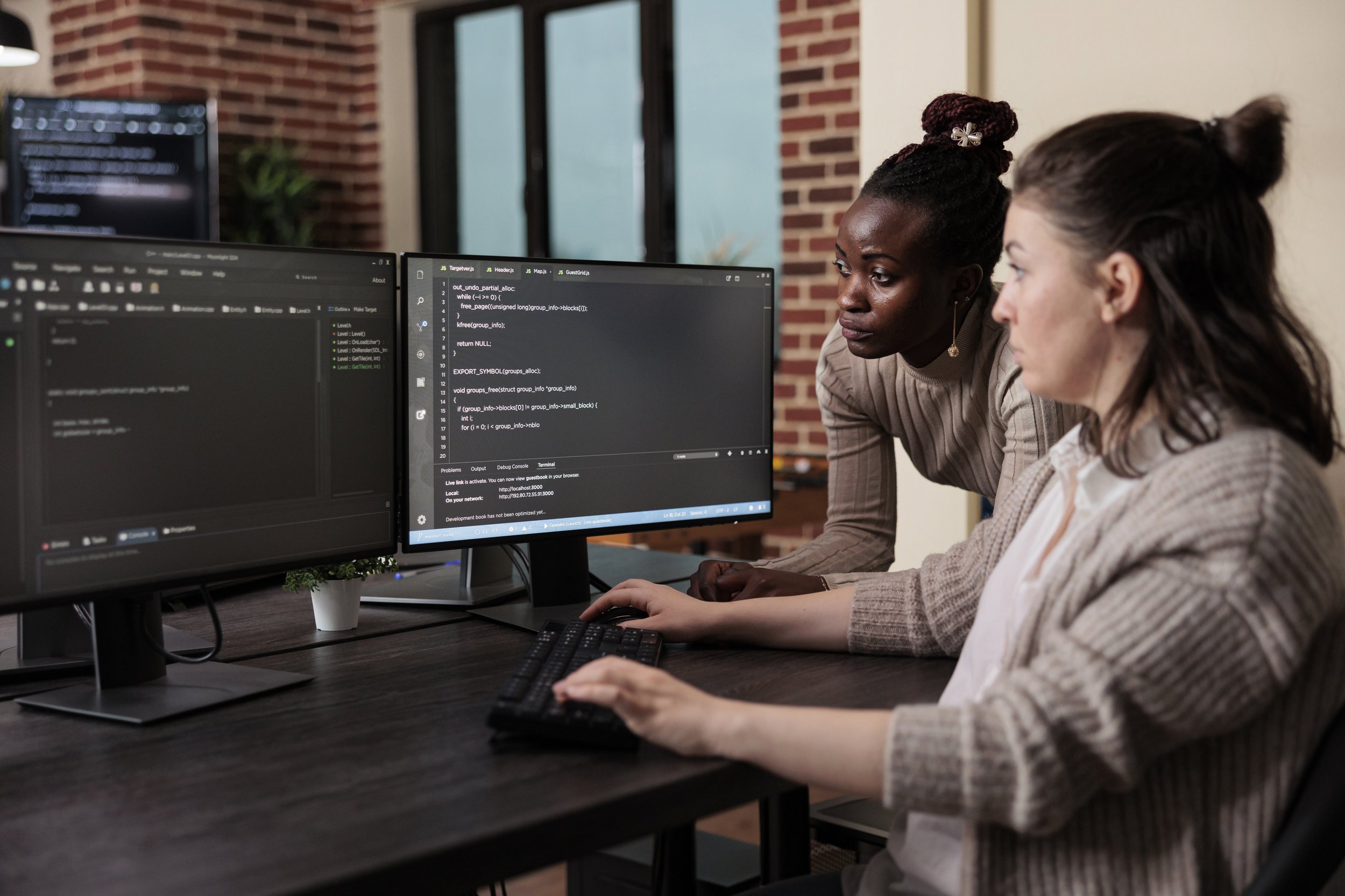 A pair of woman, one white and one Black, work on programming on a computer monitor together.