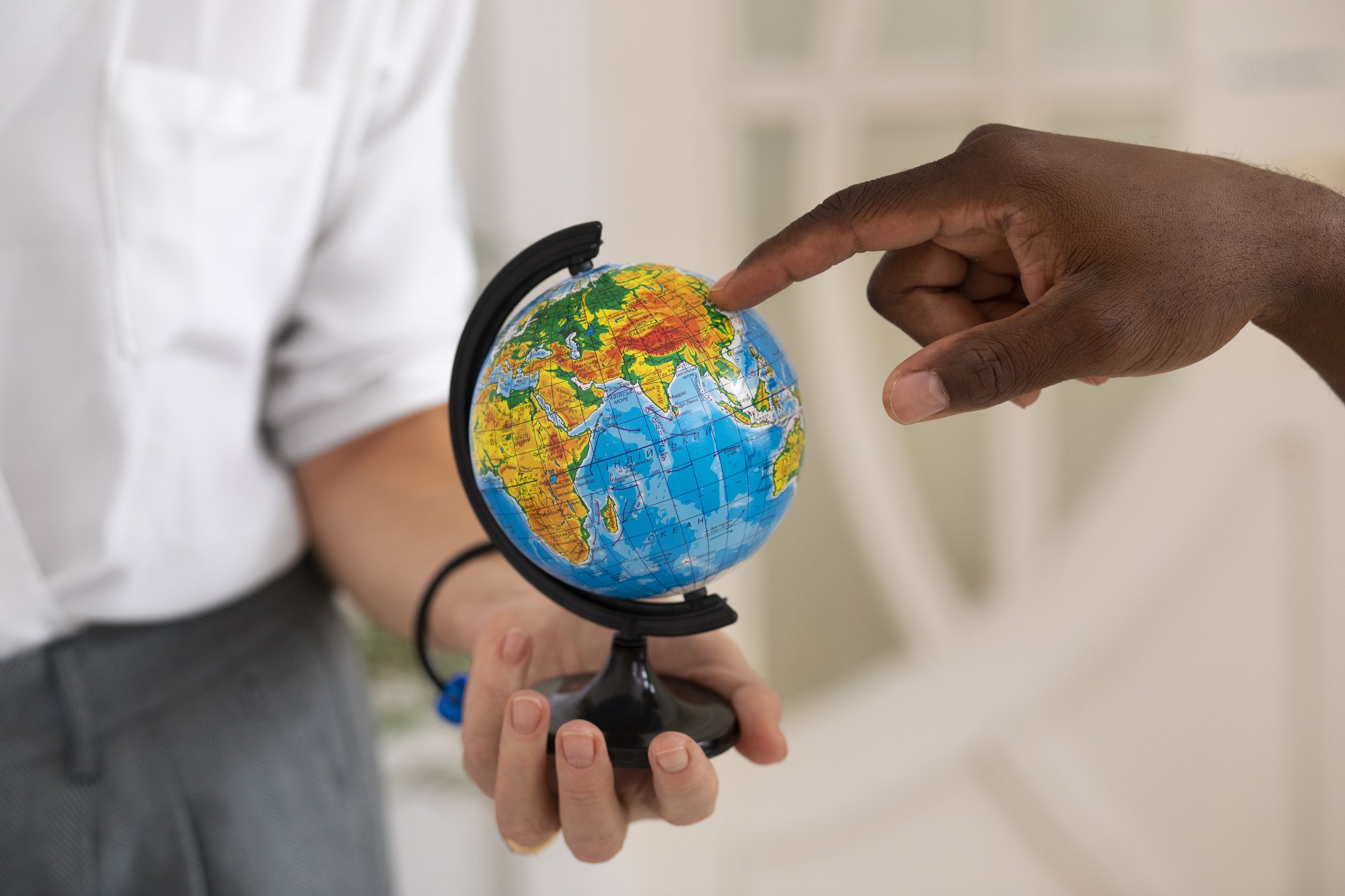 One hand holds a globe and another hand points at a spot in East Asia.