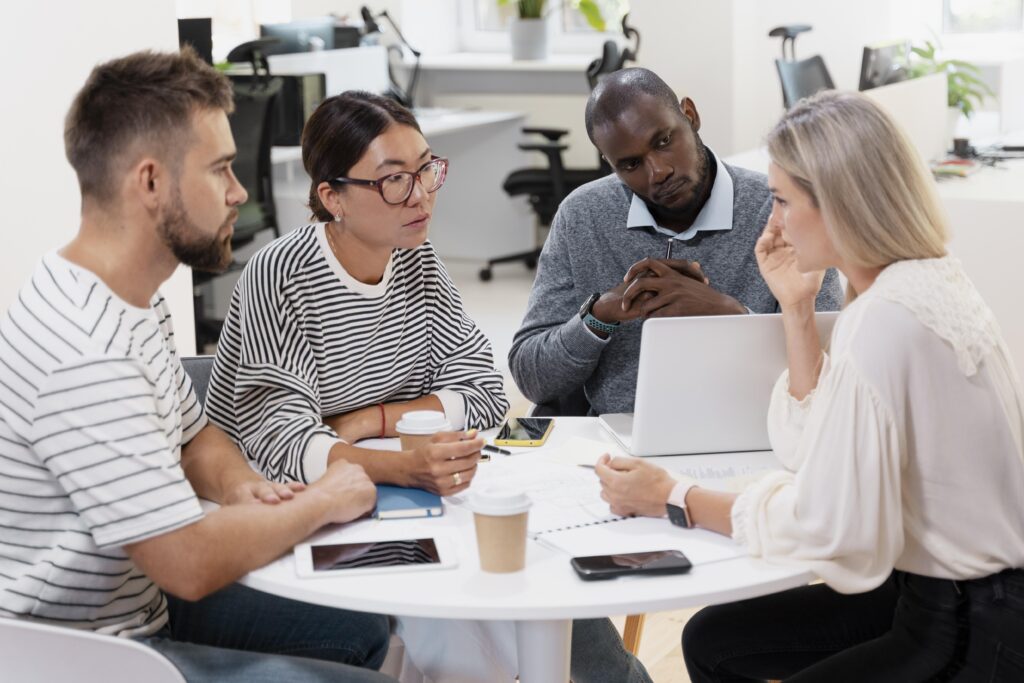 A diverse corporate team gathers in an office for a meeting and sits around a table together.