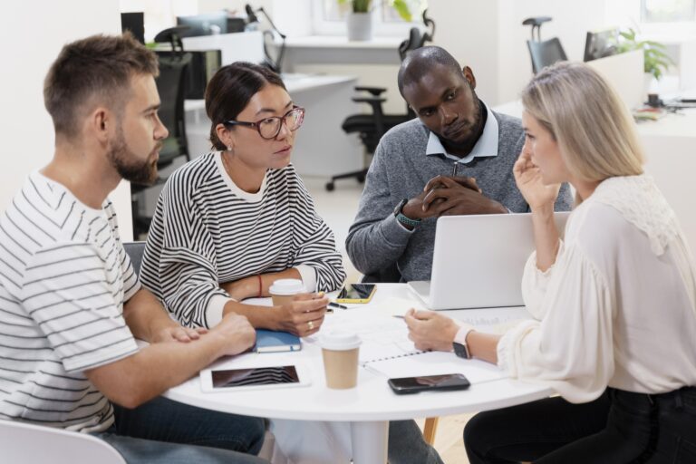 A diverse corporate team gathers in an office for a meeting and sits around a table together.