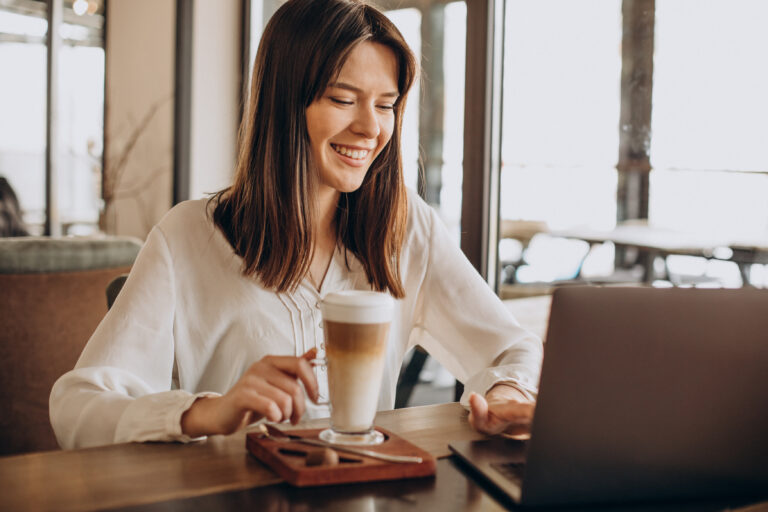 A young professional woman smiles and types on laptop in a coffee shop.