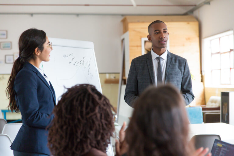 Two young corporate trainers present to a group and stand in front of a whiteboard easel.