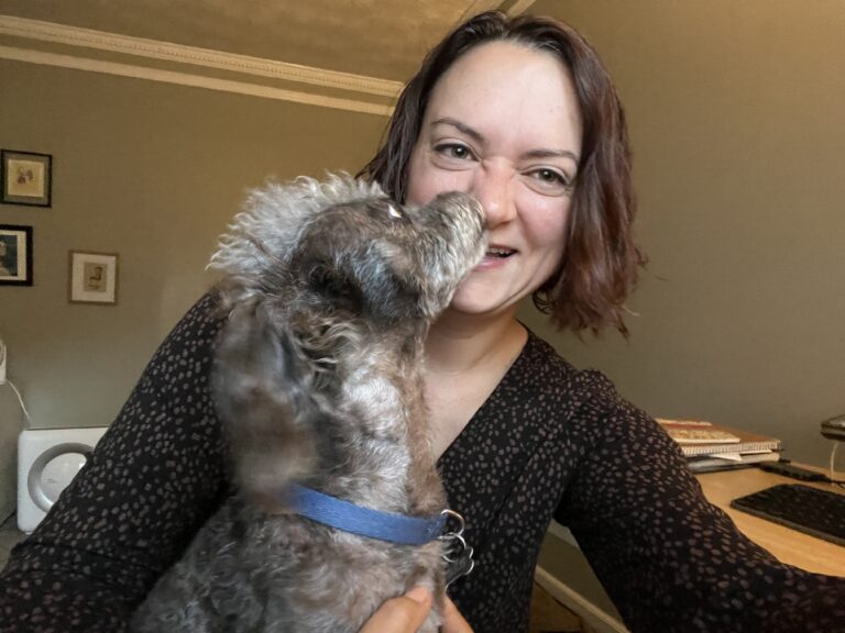 The author tries to smile for a photo at her home office desk while a dog leaps up to lick her face.