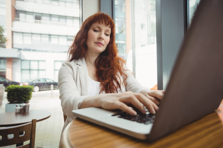 A white woman with red hair sits at a table and types on her laptop at a round cafe table.