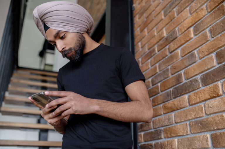 A man in a turban and black tee shirt stops in a stairwell to look at his phone.