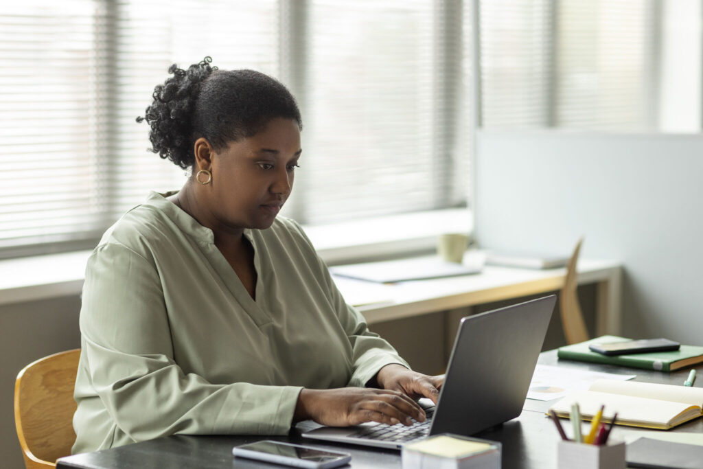 A Black woman in a light green top types at a laptop in a bright office space.