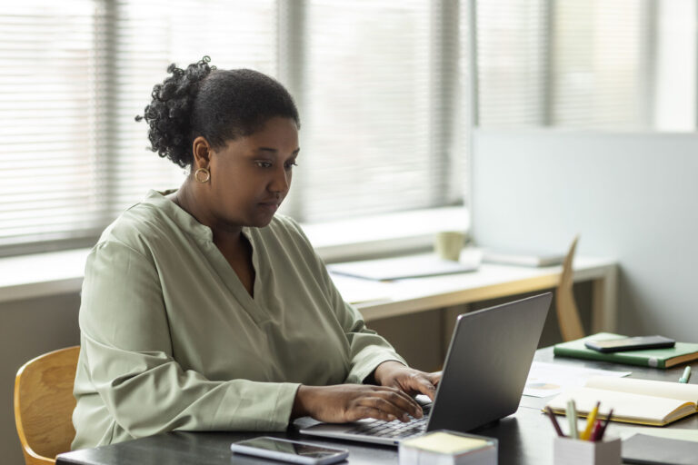 A Black woman in a light green top types at a laptop in a bright office space.
