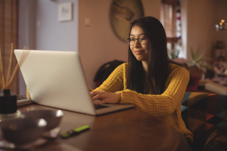 A young Asian woman uses her laptop at her dining room table.