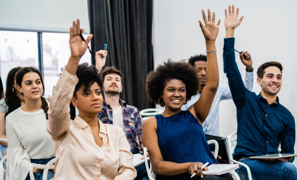 A group of young people in a corporate session face the speaker and raise their hands.