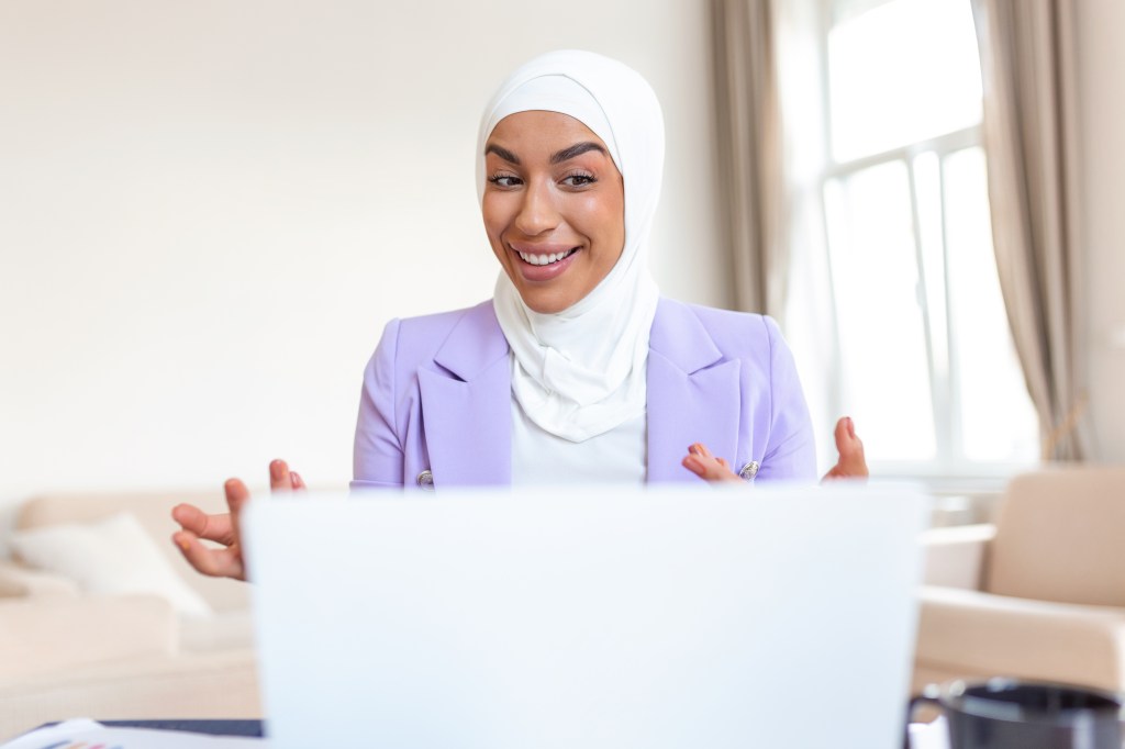 A smiling woman in a hijab talks to an audience on her laptop.