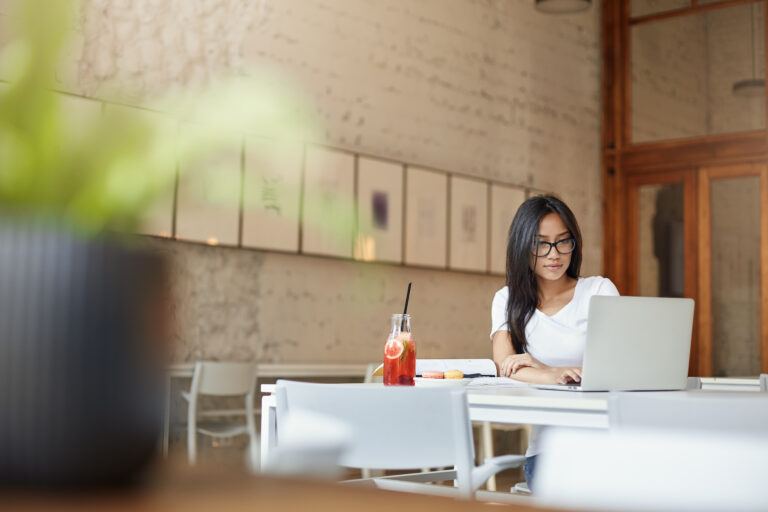A young Asian woman works at a table in a big open air cafe with a laptop and juice.