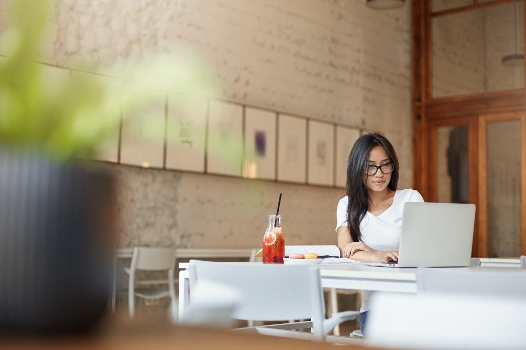 A young Asian woman works at a table in a big open air cafe with a laptop and juice.
