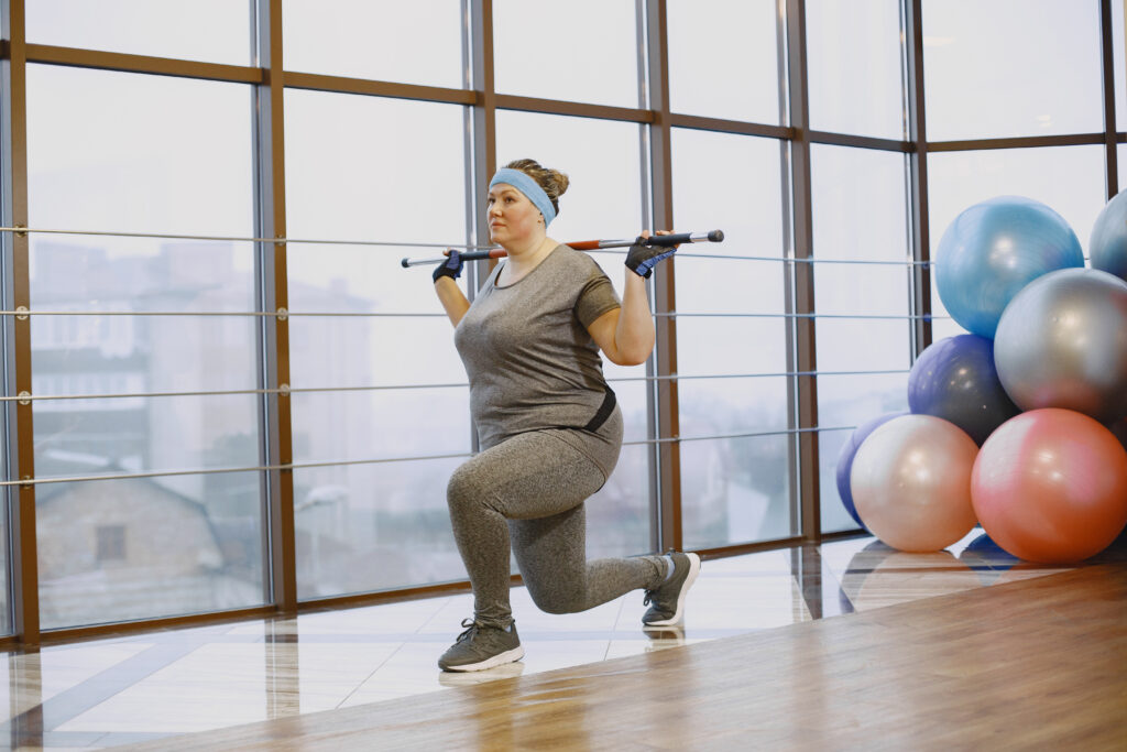 A white woman in a grey tracksuit does lunges in a gym space.