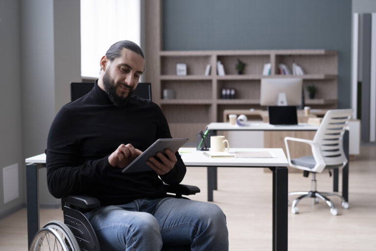 A bearded man in a wheelchair sits in his office, working on a tablet near his desk.