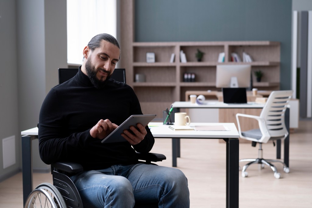 A bearded man in a wheelchair sits in his office, working on a tablet near his desk.