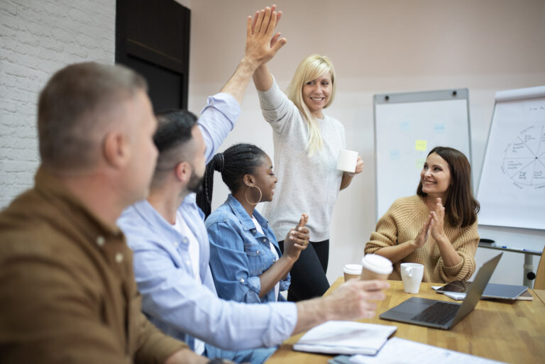 A woman standing in a board room of employees high-fives a man sitting down