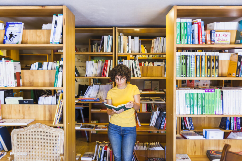 A white woman with short curly brown hair and a bright yellow t-shirt stands between bookshelves at a library looking at the open pages of a book.