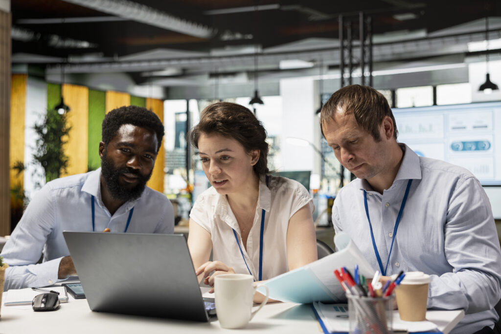 Three colleagues in an office gather around a laptop to review something together.