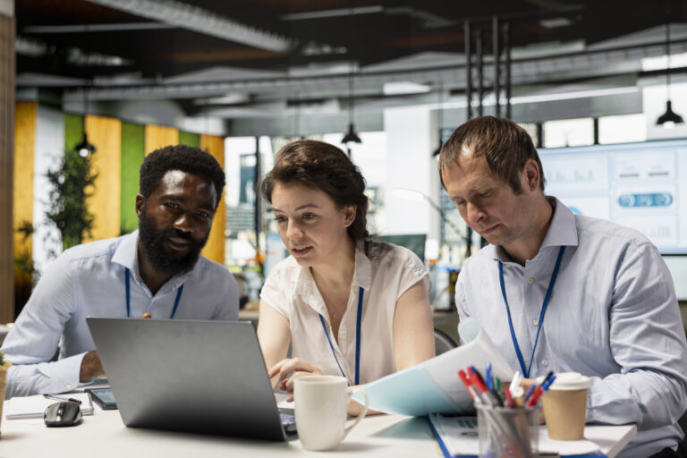 Three colleagues in an office gather around a laptop to review something together.