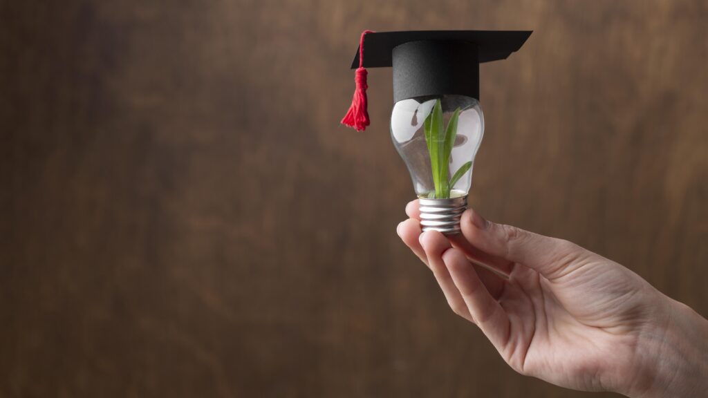 A hand holds up a small lightbulb with a graduation hat on top and a plant inside.