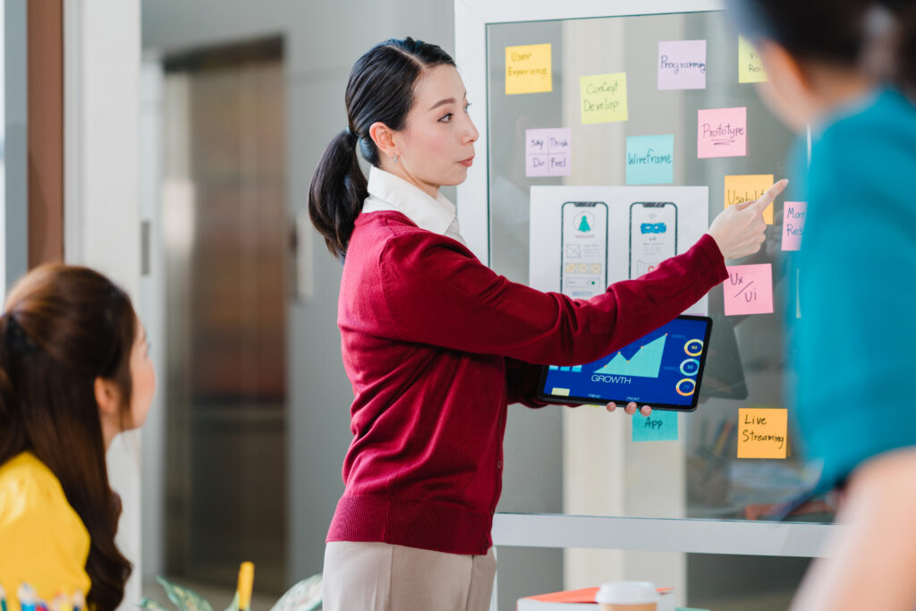 An Asian woman in a corporate office stands at a dry erase board with post-it notes set up as a Kanban board
