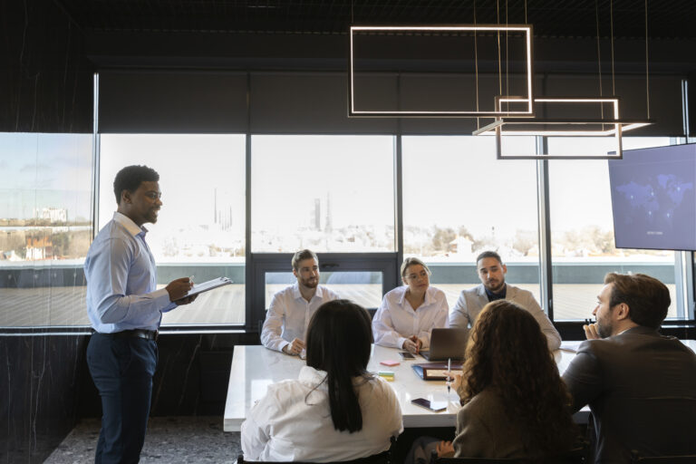 A Black male office worker stands and presents to a conference room of colleagues.