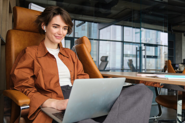 A young white woman with short brown hair and an orange jacket casually reclines in an office chair with a laptop on her legs.