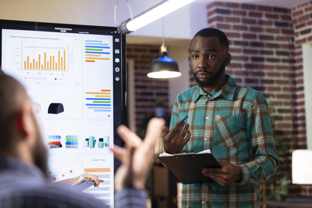 A young Black man presents a display with charts and tables in an office setting.
