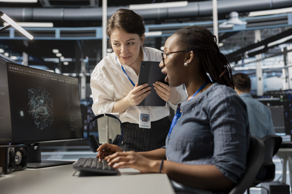 A Black woman with glasses explains a visualization on her screen to a white woman standing nearby.