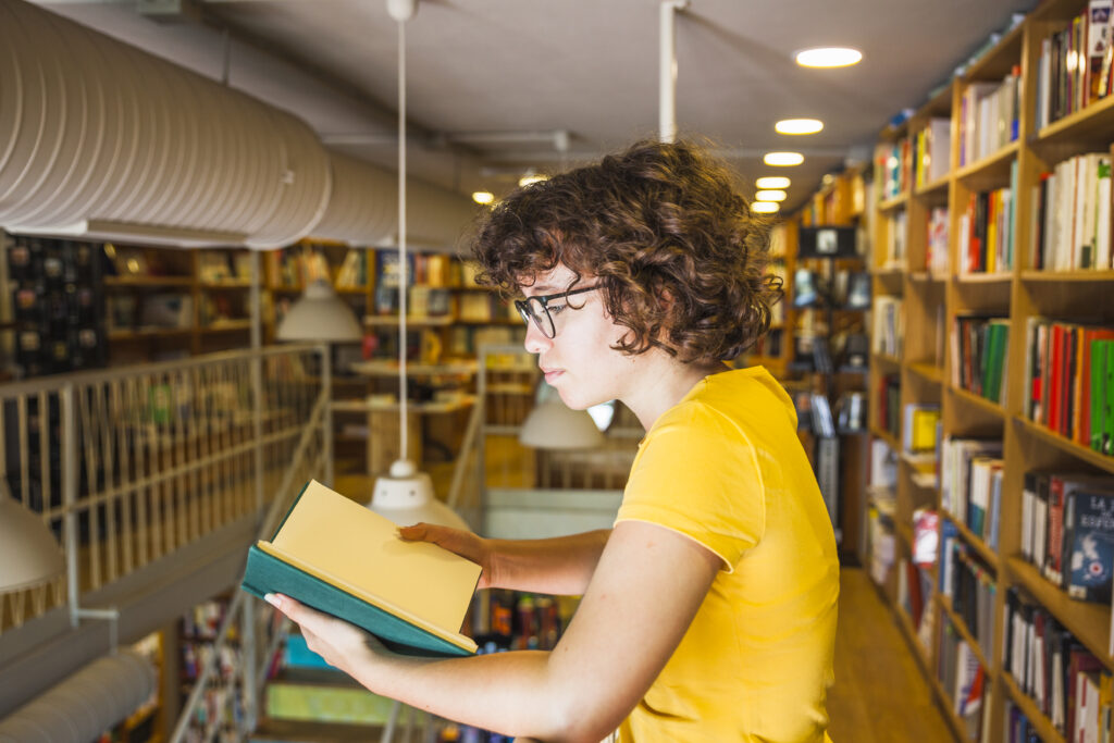 A white woman with curly brown hair and a bright yellow t-shirt reads a book in a bookstore