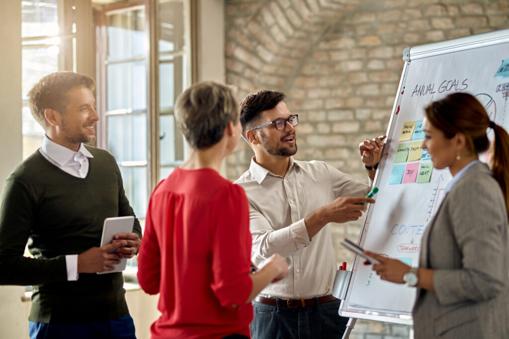 A project manager who is a man with a white button down shirt and glasses gathers an office team around a board that says Annual Goals