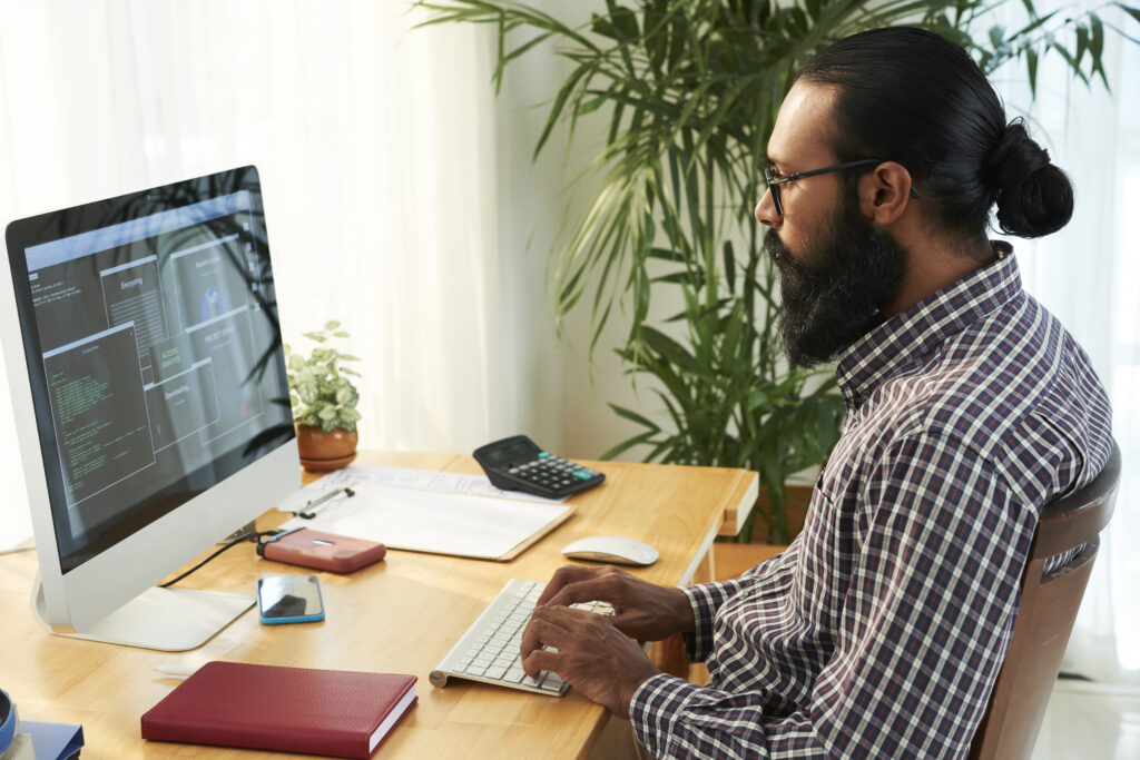 An Indian man with glasses, a plaid collared shirt, beard, and long hair in a bun sits at a desk typing into a code editor on his monitor.